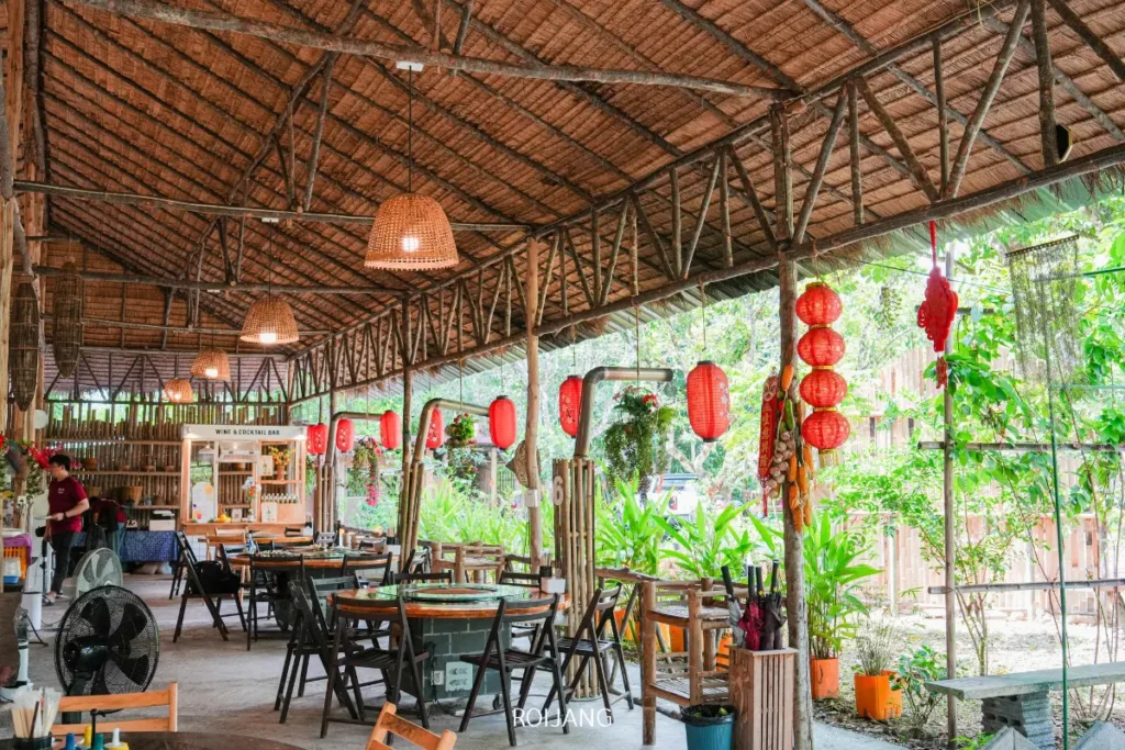 ร้านอาหารกลางแจ้งที่มีหลังคาทำจากไม้ไผ่ โต๊ะและเก้าอี้ไม้ โคมไฟสีแดงแขวนเป็นของตกแต่ง และต้นไม้สีเขียวสามารถมองเห็นได้จากภายนอก บางคนกำลังเพลิดเพลินกับ ไก่ไฟฟืน ในพื้นหลัง.