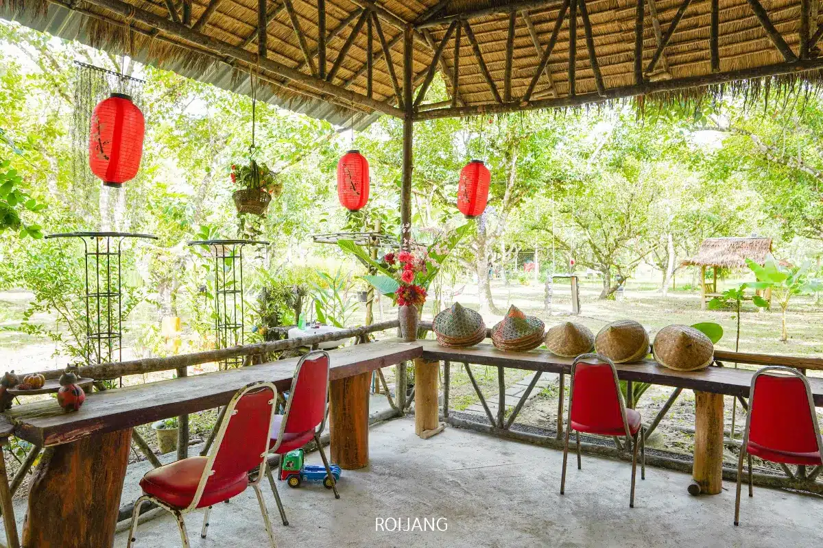 ศาลาเปิดโล่งพร้อมโคมแดง โต๊ะไม้ เก้าอี้สีแดง ตะกร้าสานจัดแสดง และพืชพรรณเขียวขจีเป็นฉากหลัง—เหมาะอย่างยิ่งสำหรับการเพลิดเพลินกับ ไก่ไฟฟืน แบบดั้งเดิมในบรรยากาศที่มีเสน่ห์.
