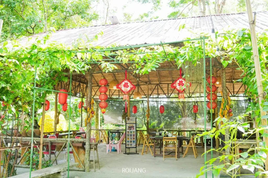 คาเฟ่กลางแจ้งที่มีหลังคาไม้ไผ่ โต๊ะและเก้าอี้ไม้ ตกแต่งด้วยโคมไฟสีแดงและต้นไม้สีเขียว เพลิดเพลินกับเมนูพิเศษ ไก่ย่างฟืน ที่ล้อมรอบด้วยพืชพรรณและต้นไม้เขียวชอุ่ม.