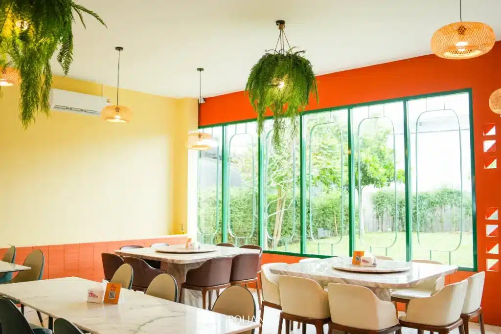 ร้านพริกกะเหรี่ยงมีบรรยากาศภายในคาเฟ่ที่สว่างสดใสและทันสมัย ประกอบด้วยโต๊ะหินอ่อน เก้าอี้หลากหลายแบบ ต้นไม้แขวน ผนังสีส้มและเหลือง และหน้าต่างบานใหญ่ที่มองเห็นวิวสีเขียว.