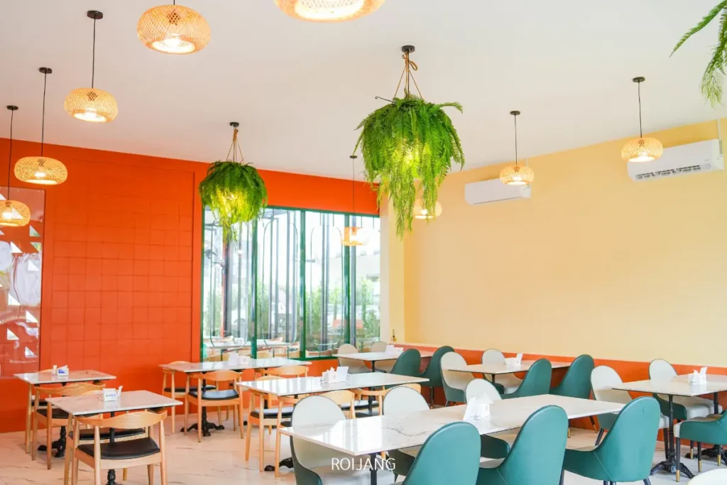 พื้นที่รับประทานอาหารในคาเฟ่สมัยใหม่ที่ร้านพริกกะเหรี่ยงมีเก้าอี้สีเขียว โต๊ะสีขาว เฟิร์นแขวน ไฟเพดานแบบแขวนที่ให้ความอบอุ่น และผนังสีส้มและเหลืองสดใส.