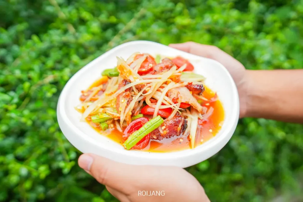 บุคคลหนึ่งถือจานขาวที่มีส้มตำไทยพร้อมมะเขือเทศ ถั่วฝักยาว และน้ำปรุงรส อยู่นอกอาคาร โดยมีใบไม้สีเขียวเป็นฉากหลัง ที่ ร้านพริกกะเหรี่ยง.