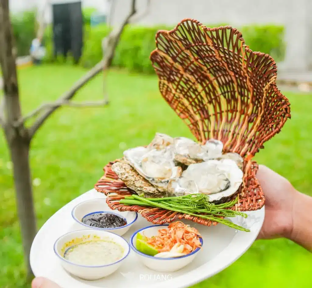 มือหนึ่งถือจานที่มีตะกร้าสานรูปเปลือกหอยบรรจุหอยนางรมจาก ร้านพริกกะเหรี่ยง เสิร์ฟพร้อมชามเครื่องปรุงและเครื่องเคียงขนาดเล็กสามใบ วางกลางแจ้งท่ามกลางหญ้าและต้นไม้เป็นฉากหลัง.