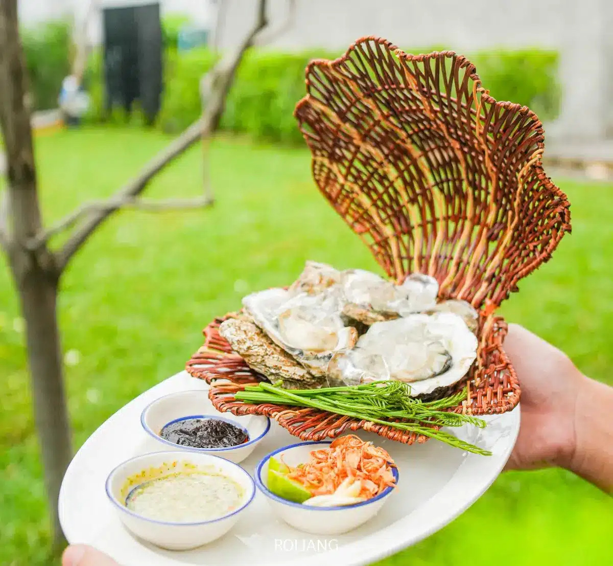 มือหนึ่งถือจานที่มีตะกร้าสานรูปเปลือกหอยบรรจุหอยนางรมจาก ร้านพริกกะเหรี่ยง เสิร์ฟพร้อมชามเครื่องปรุงและเครื่องเคียงขนาดเล็กสามใบ วางกลางแจ้งท่ามกลางหญ้าและต้นไม้เป็นฉากหลัง.