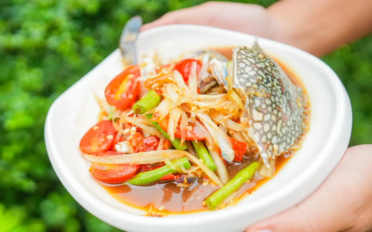 ชามสีขาวใส่อาหารเป็นส้มตำไทยรสเผ็ด มีมะเขือเทศ ถั่วฝักยาว พริก และปูม้าดิบ เสิร์ฟโดย ร้านพริกกะเหรี่ยง และถือด้วยสองมือกลางแจ้ง.