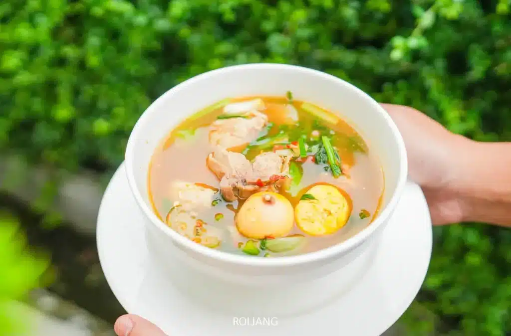 มือหนึ่งถือชามซุปสีขาวที่มีไก่ ไข่ไก่เล็ก สมุนไพร และผักจาก ร้านพริกกะเหรี่ยง วางไว้บนพื้นหลังสีเขียวที่เบลอ.
