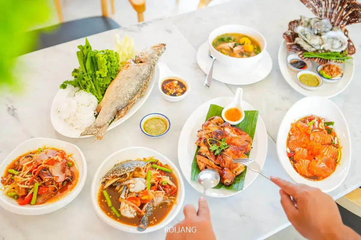 โต๊ะที่ร้านพริกกะเหรี่ยงมีอาหารไทยเช่น ปลาเผา, ไก่ย่าง, ซุป, หอยนางรม, สลัด, และผักสด, พร้อมไก่เสิร์ฟด้วยมือ.