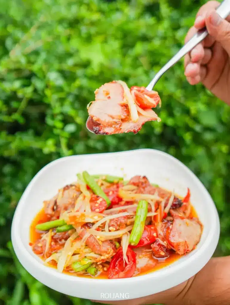 มือหนึ่งถือช้อนที่มีหมูหั่นบางวางอยู่เหนือชามสลัดเผ็ดที่มีผัก มะเขือเทศ และถั่วฝักยาว ร้านพริกกะเหรี่ยง ฉากหลังเป็นพืชสีเขียวสดใส.