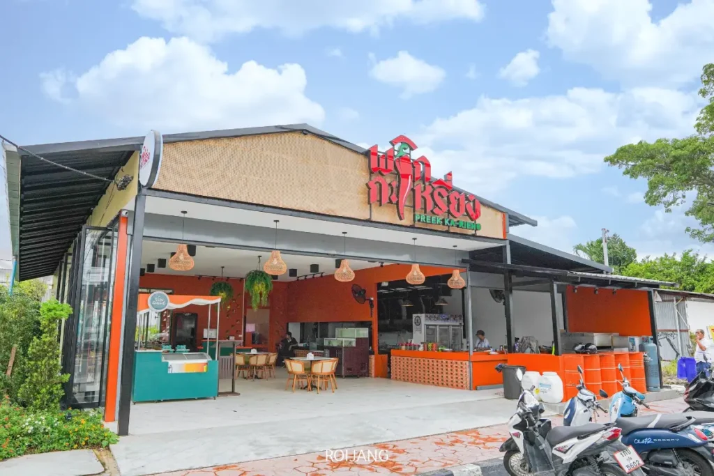 ร้านอาหารสมัยใหม่เปิดด้านหน้า ตกแต่งด้วยสีส้มและไม้ มีที่นั่งกลางแจ้ง มีสกู๊ตเตอร์จอดอยู่ด้านหน้า และมีป้ายเขียนว่า "พริกกะเหรี่ยง" (ร้านพริกกะเหรี่ยง) เป็นภาษาไทยและภาษาอังกฤษ.