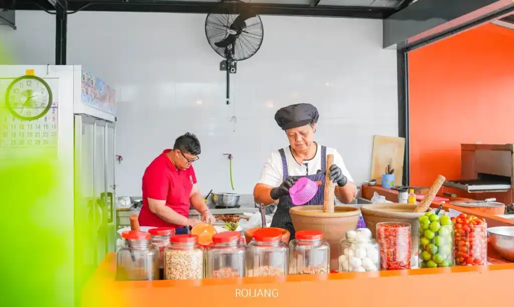 สองคนกำลังเตรียมอาหารในครัวที่สว่างไสว มีขวดโหลของส่วนผสมจาก ร้านพริกกะเหรี่ยง บนเคาน์เตอร์; คนหนึ่งกำลังหั่นผัก ในขณะที่อีกคนหนึ่งใช้ครกและสาก.