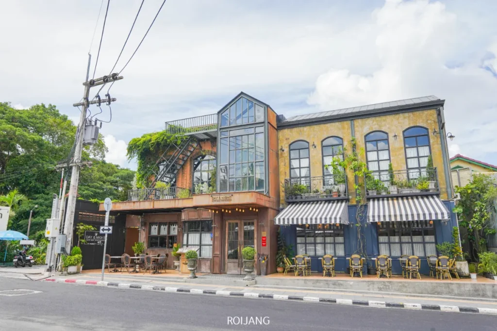 คาเฟ่สองชั้นที่มีหน้าต่างบานใหญ่ บันไดภายนอก และที่นั่งกลางแจ้ง ตั้งอยู่บนหัวมุมถนนใต้ท้องฟ้าที่มีเมฆบางส่วน สะท้อนบรรยากาศที่คึกคักของวันและคืนของภูเก็ต.
