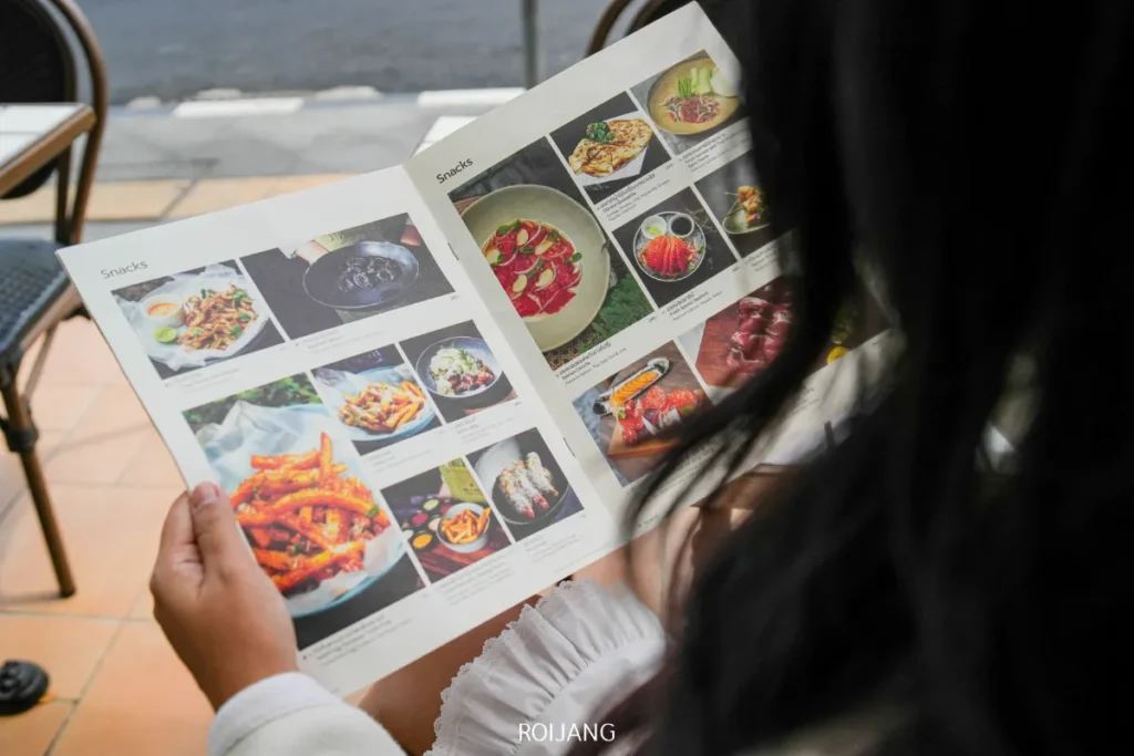 บุคคลกำลังถือและดูเมนูร้านอาหาร "เดย์ แอนด์ ไนท์" ของภูเก็ต ซึ่งมีรูปภาพและคำอธิบายของอาหารหลากหลายชนิด นั่งอยู่ที่โต๊ะกลางแจ้ง.