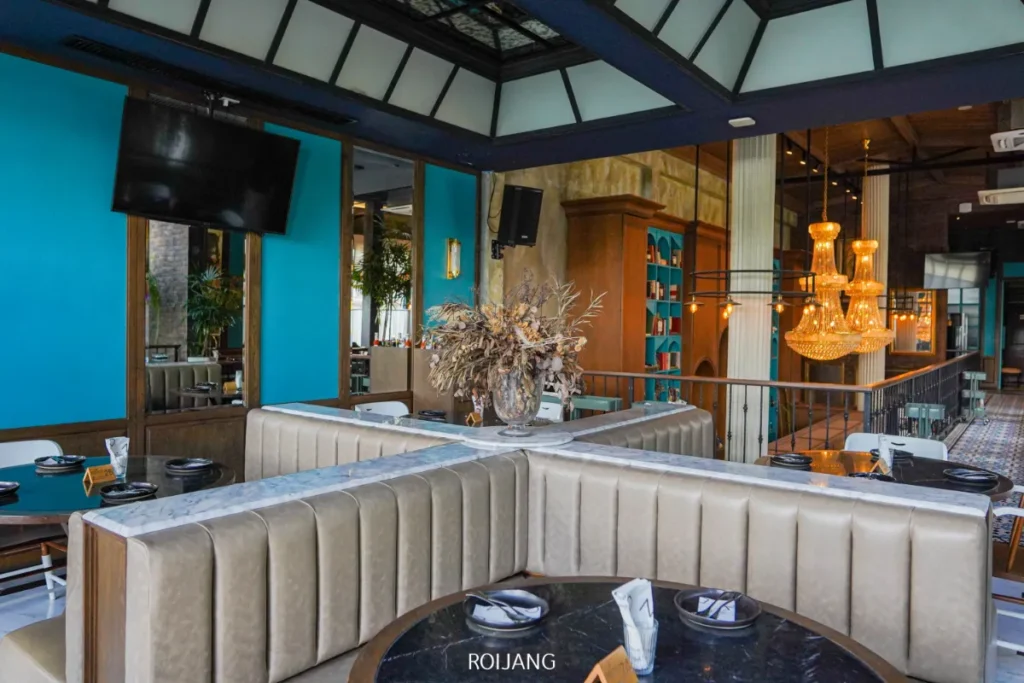 ร้านอาหารสมัยใหม่ที่มีบรรยากาศภายในตกแต่งด้วยที่นั่งแบบบูธสีเบจ โต๊ะกลมที่จัดวางจานและผ้าเช็ดปากไว้เรียบร้อย ผนังสีฟ้า โคมไฟระย้า และแจกันดอกไม้แห้งขนาดใหญ่เป็นจุดเด่นตรงกลาง—ถ่ายทอดบรรยากาศสุดชิคของ Day & Night แห่งภูเก็ต.