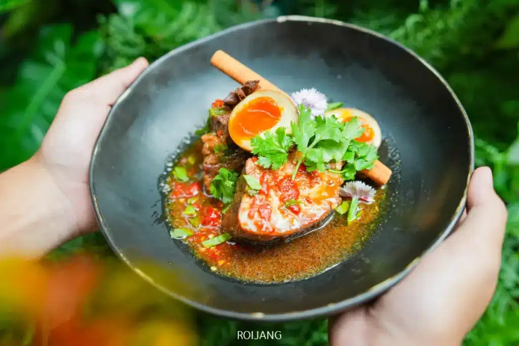 บุคคลหนึ่งถือชามสีดำที่มีอาหารจานปลาเผ็ดจากร้านวัน & คืนของภูเก็ต โรยด้วยสมุนไพร พริกแดงหั่นแว่น และไข่ลวกผ่าครึ่งในน้ำซุปสีสดใส.