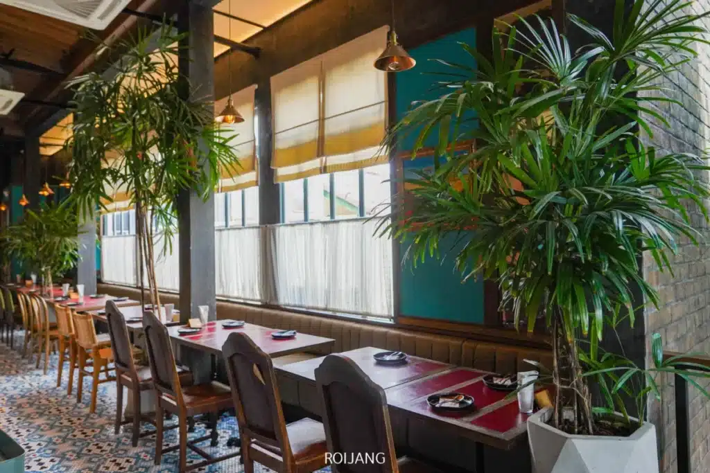 ร้านอาหารสมัยใหม่ภายใน Day & Night of Phuket โดดเด่นด้วยต้นไม้กระถางขนาดใหญ่ โต๊ะไม้ที่จัดวางด้วยแผ่นรองจานและเครื่องใช้บนโต๊ะอาหาร พร้อมแสงธรรมชาติที่ส่องผ่านหน้าต่างบานกว้าง.