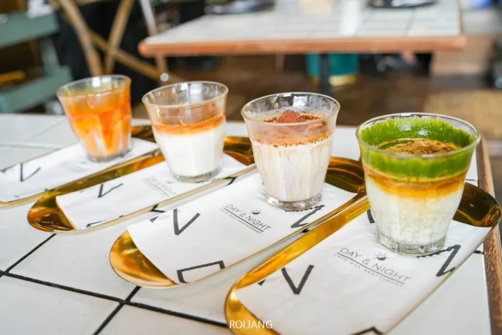 ขนมหวานสี่แก้วที่จัดเป็นชั้นๆ แต่ละชั้นมีท็อปปิ้งที่แตกต่างกัน เสิร์ฟบนถาดทองคำพร้อมด้วยผ้าเช็ดปากที่มีแบรนด์ Day & Night of Phuket บนโต๊ะกระเบื้องในบรรยากาศคาเฟ่ที่อบอุ่น.