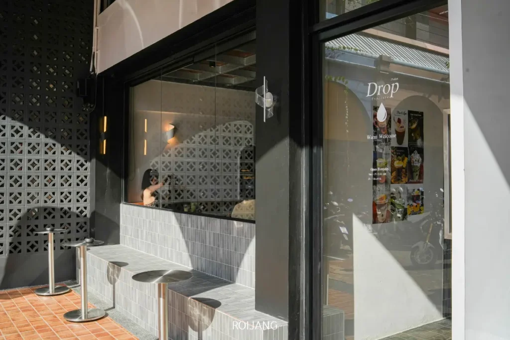 พื้นที่นั่งเล่นกลางแจ้งของ Pause Coffee Bar มีม้านั่งปูกระเบื้องและเก้าอี้ทรงกลม หน้าต่างบานใหญ่ และเมนูที่แสดงบนประตูกระจก.
