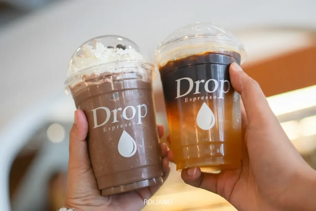 สองมือถือแก้วพลาสติกใส่น้ำแข็งจาก Drop Espresso และ Pause Coffee Bar—หนึ่งแก้วช็อกโกแลตกับวิปครีม และหนึ่งแก้วกาแฟดำ ทั้งสองแก้วมีฝาโดมปิดอยู่ด้านบน.