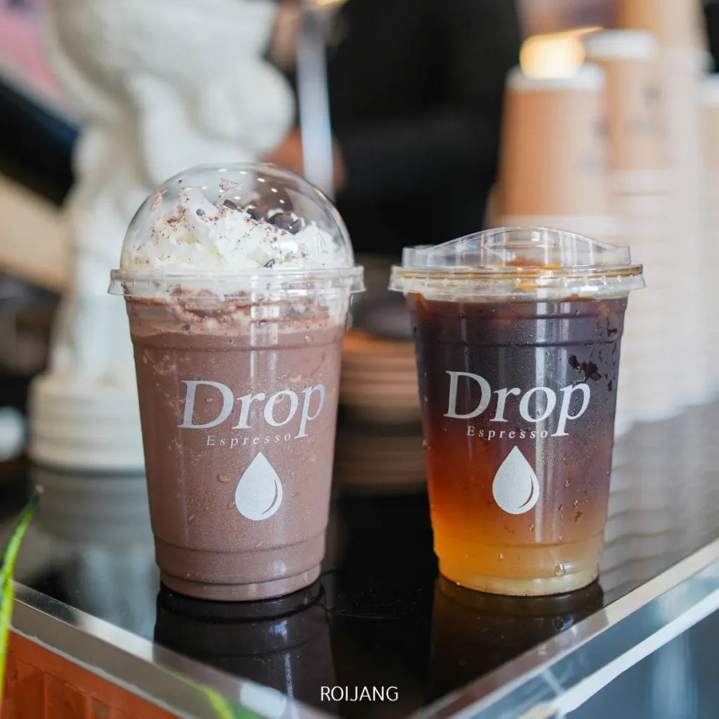 เครื่องดื่มสองแก้วจาก Drop Espresso—ช็อกโกแลตเย็นกับวิปครีมและกาแฟดำเย็น—วางอยู่บนเคาน์เตอร์สะท้อนแสงที่ Pause Coffee Bar.