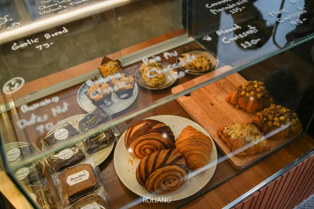 ตู้โชว์ขนมที่ Pause Coffee Bar จัดแสดงครัวซองต์ บราวนี่ มัฟฟิน และขนมปังบนจานและถาด โดยมีชื่อและราคาของสินค้าเขียนไว้บนกระจก.