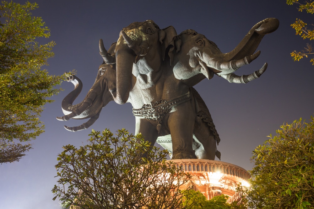 รูปปั้นช้างสามเศียรขนาดใหญ่ตั้งอยู่บนแท่นที่ตกแต่งอย่างสวยงาม ส่องสว่างในยามค่ำคืนด้วยต้นไม้ในเบื้องหน้า เป็นภาพที่น่าตื่นตาตื่นใจสำหรับผู้ที่มองหาสถานที่ท่องเที่ยวบางปูและสถานที่ท่องเที่ยวทางวัฒนธรรมในบริเวณใกล้เคียง.