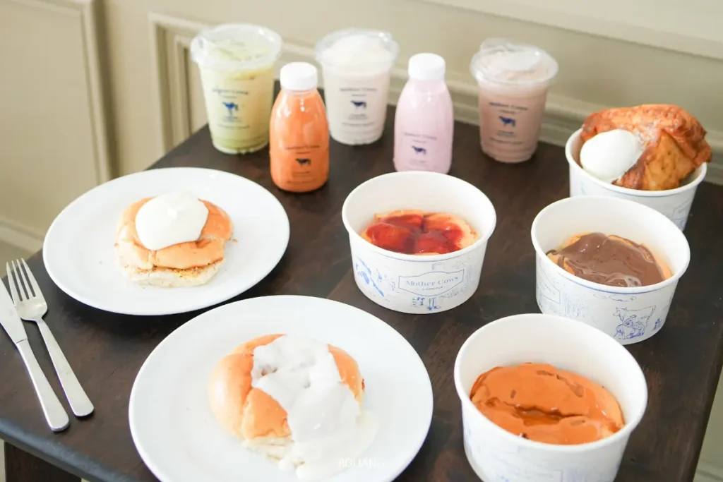ขนมหวานและเครื่องดื่มหลากหลายจาก Mother Cows เขาหลัก บนโต๊ะไม้สีเข้ม ประกอบด้วยขนมปังกับครีม ถ้วยพุดดิ้ง เครื่องดื่มบรรจุขวด และถ้วยเยลลี่กับคัสตาร์ด.