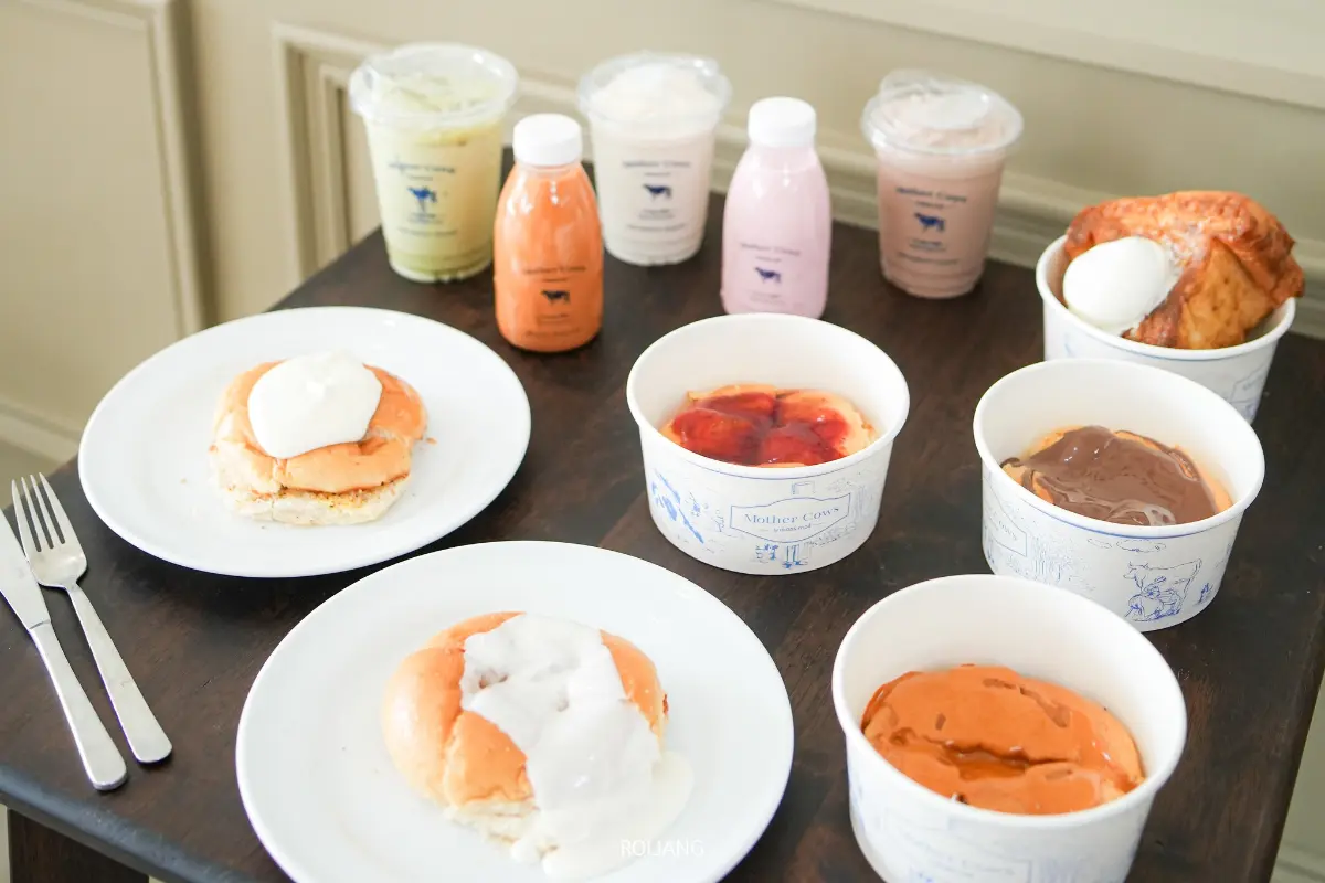 ขนมหวานและเครื่องดื่มหลากหลายจาก Mother Cows เขาหลัก บนโต๊ะไม้สีเข้ม ประกอบด้วยขนมปังกับครีม ถ้วยพุดดิ้ง เครื่องดื่มบรรจุขวด และถ้วยเยลลี่กับคัสตาร์ด.