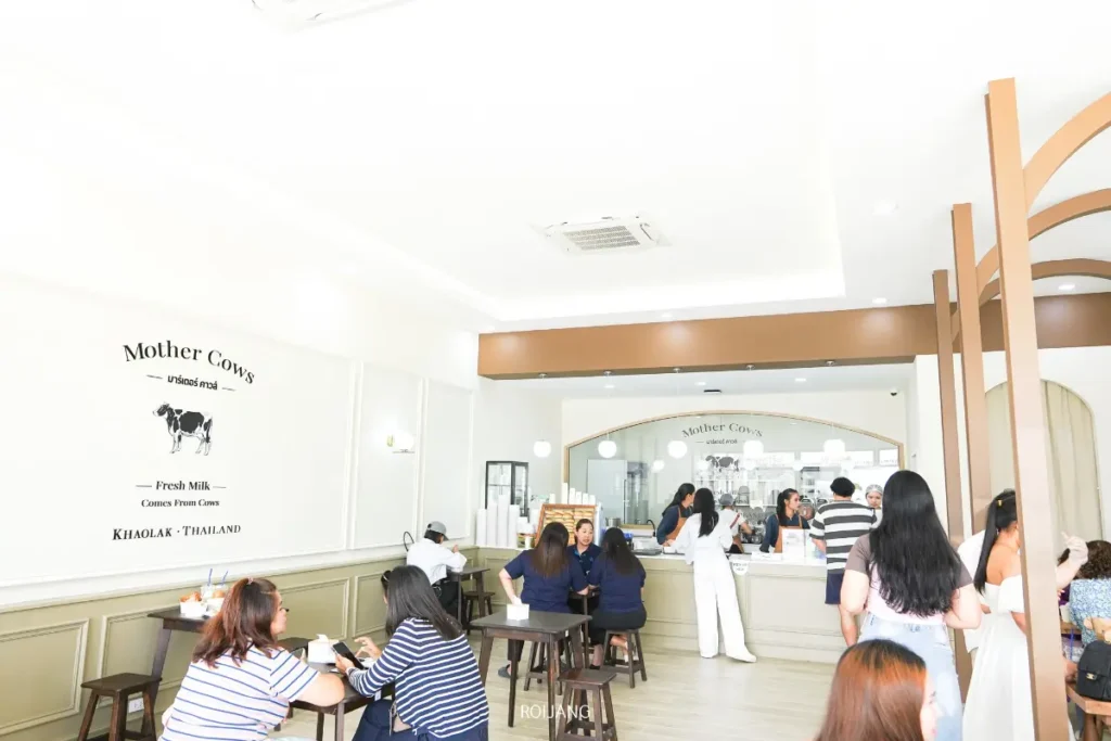 ภายในคาเฟ่ที่สว่างและทันสมัย มีผู้คนหลายนั่งอยู่ที่โต๊ะและบางคนยืนอยู่ใกล้เคาน์เตอร์ใต้ป้าย "แม่วัว เขาหลัก" บนผนัง.