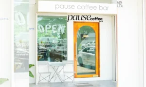 คาเฟ่เล็กๆ ที่มีประตูกระจก เก้าอี้โลหะสองตัว และโต๊ะหนึ่งตัวด้านนอก ด้านบนของทางเข้า ป้าย Pause Coffee Bar ต้อนรับผู้มาเยือน หน้าต่างแสดงคำว่า "เปิด" และ "Depresso.