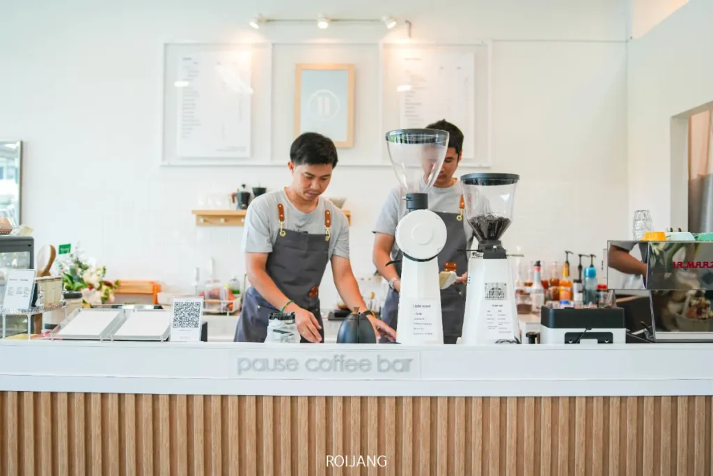 บาริสต้าสองคนทำงานอยู่หลังเคาน์เตอร์ที่ Pause Coffee Bar โดยเชี่ยวชาญในการเตรียมเครื่องดื่มด้วยเครื่องชงกาแฟและอุปกรณ์สมัยใหม่.