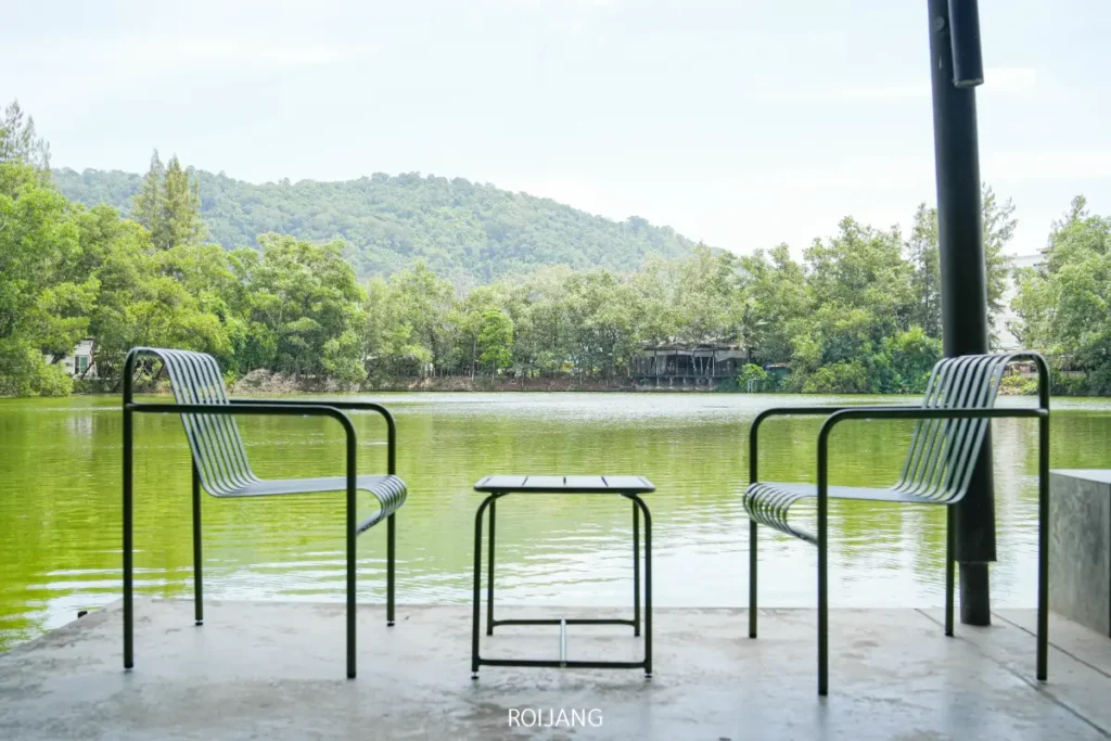 เก้าอี้โลหะสองตัวและโต๊ะเล็กหนึ่งตัวจาก Pause Coffee Bar หันหน้าไปทางทะเลสาบสีเขียว โดยมีต้นไม้และเนินเขาเป็นฉากหลัง มองเห็นจากลานคอนกรีต.