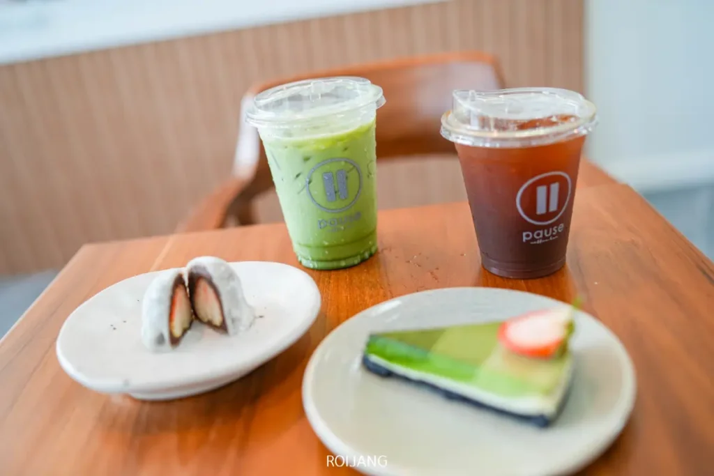 ที่ Pause Coffee Bar แก้วพลาสติกสองใบพร้อมเครื่องดื่ม, ชิ้นเค้กสีเขียวกับสตรอเบอร์รี่, และของหวานโมจิครึ่งชิ้นบนจานสีขาว ถูกจัดวางอย่างสวยงามบนโต๊ะไม้.