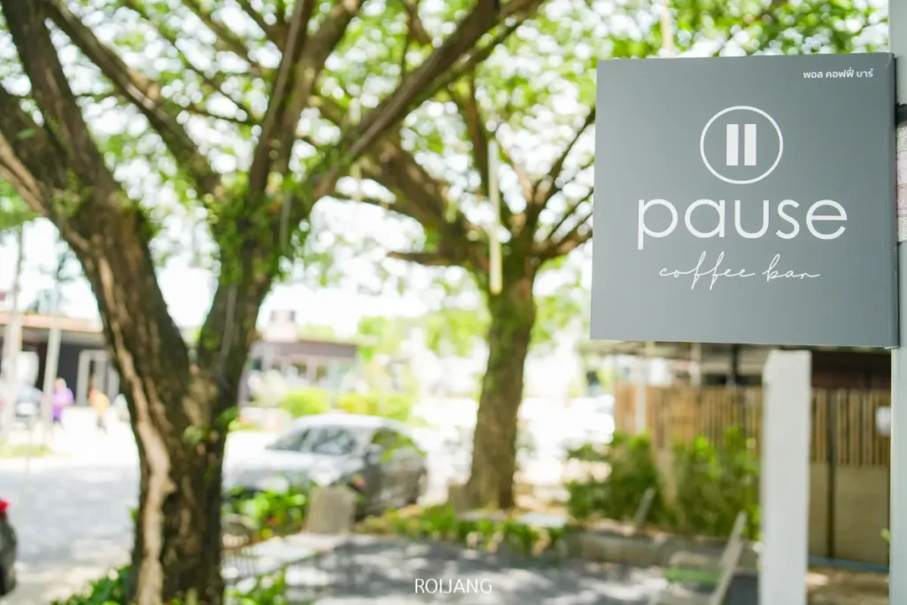ป้ายสีเทาที่เขียนว่า "Pause Coffee Bar" แขวนอยู่ด้านนอก โดยมีต้นไม้ใบเขียวและรถที่จอดอยู่เป็นฉากหลัง.