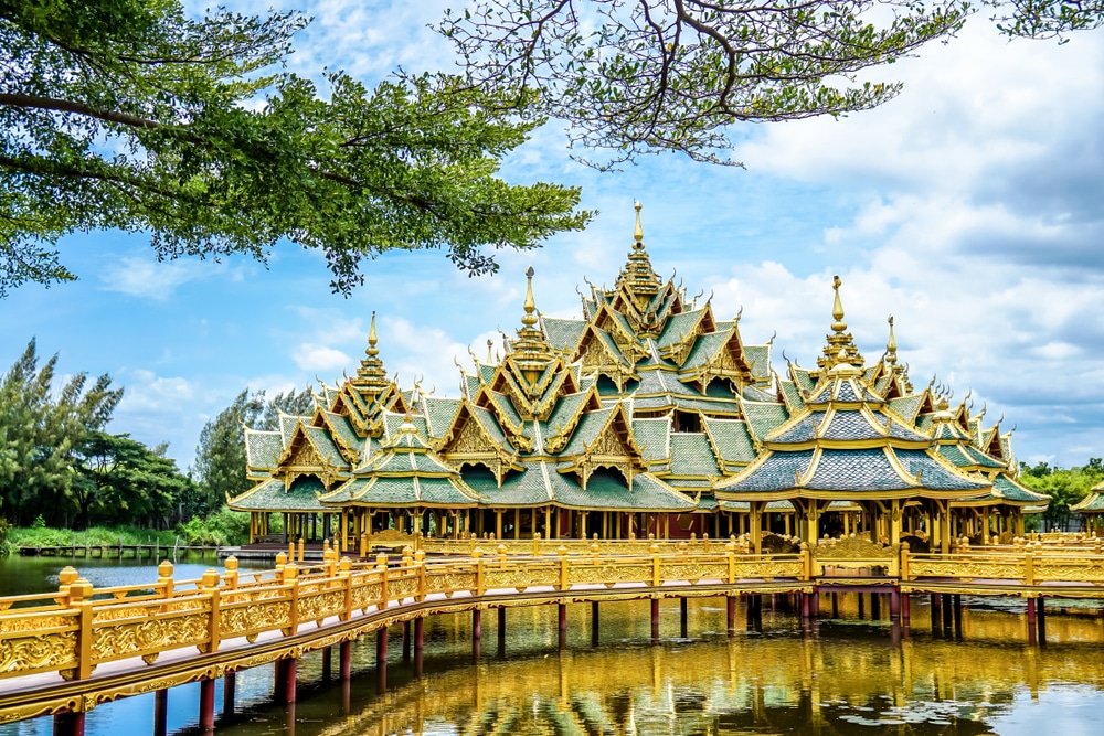 ศาลาไทยแบบดั้งเดิมที่มีหลังคาทองคำประดับอย่างสวยงามตั้งอยู่เหนือสระน้ำที่สะท้อนเงา ที่บางปูที่เที่ยว ล้อมรอบด้วยต้นไม้เขียวชอุ่มและต้นไม้ใหญ่ภายใต้ท้องฟ้าที่มีเมฆบางส่วน.