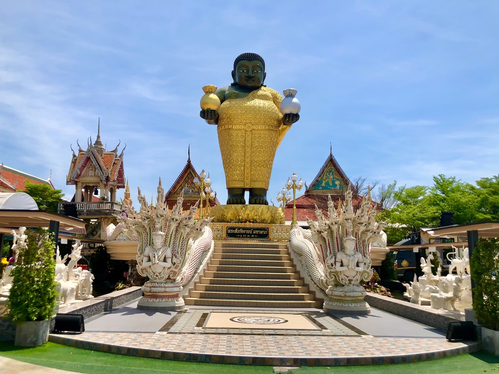 พระพุทธรูปทองคำขนาดใหญ่ถือวัตถุสองชิ้นตั้งอยู่บนแท่นที่ตกแต่งอย่างสวยงาม ขนาบข้างด้วยประติมากรรมสีขาวที่ประดับประดาและอาคารวัดภายใต้ท้องฟ้าสีคราม—เป็นจุดเด่นที่น่าประทับใจสำหรับผู้ที่มาเยือนสถานที่ท่องเที่ยวบางปู.