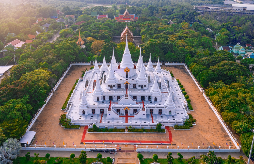 ภาพมุมสูงของวัดสีขาวขนาดใหญ่ที่มีเจดีย์หลายยอด ล้อมรอบด้วยต้นไม้สีเขียวและทางเดินในลานกว้าง—เหมาะสำหรับผู้ที่กำลังมองหาสถานที่ท่องเที่ยวบางปู หรือวางแผนที่จะเที่ยวบางปู.