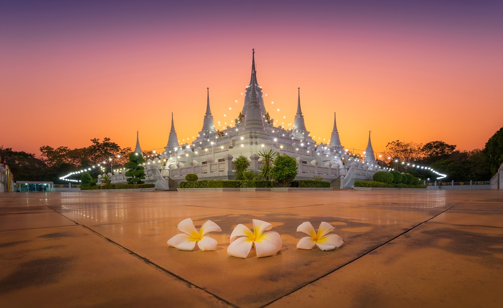 วัดขาวที่มียอดแหลมหลายยอด ส่องสว่างด้วยไฟประดับเมื่อพระอาทิตย์ตกดิน พร้อมดอกจำปีสีขาวโปรยปรายอยู่บนพื้น—เป็นภาพที่สงบเงียบเหมาะสำหรับผู้ที่กำลังค้นหา ที่เที่ยวบางปู และช่วงเวลาที่สงบใกล้กับบางปู.