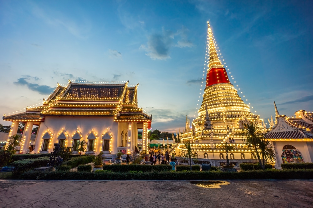 วัดและเจดีย์ไทยประดับด้วยไฟประดับสีสันสดใสในยามพลบค่ำ มีผู้คนเดินเล่นรอบบริเวณและท้องฟ้าที่มีเมฆบางส่วน—เป็นภาพที่งดงามน่าหลงใหลที่ทำให้ บางปูที่เที่ยว เป็นสถานที่ที่เหมาะสำหรับการเยี่ยมชมในตอนเย็น.