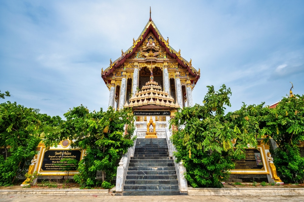 วัดไทยแบบดั้งเดิมที่มีรายละเอียดสีทองและสีแดงอันวิจิตร บันไดหิน และต้นไม้เขียวขจีทั้งสองข้างใต้ท้องฟ้าสีคราม—เป็นสถานที่เงียบสงบที่ชวนให้นึกถึงบรรยากาศอันสงบสุขที่ ที่เที่ยวบางปู.