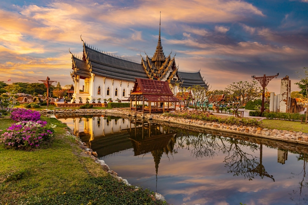 วัดไทยแบบดั้งเดิมที่มีหลังคาประดับอย่างวิจิตรตั้งอยู่ข้างสระน้ำที่สะท้อนภาพอาคาร ล้อมรอบด้วยสวนดอกไม้ภายใต้ท้องฟ้าสีสันสวยงามยามพระอาทิตย์ตกดิน—เหมาะสำหรับผู้ที่ต้องการสถานที่ท่องเที่ยวบางปู และการพักผ่อนที่สงบเงียบ.