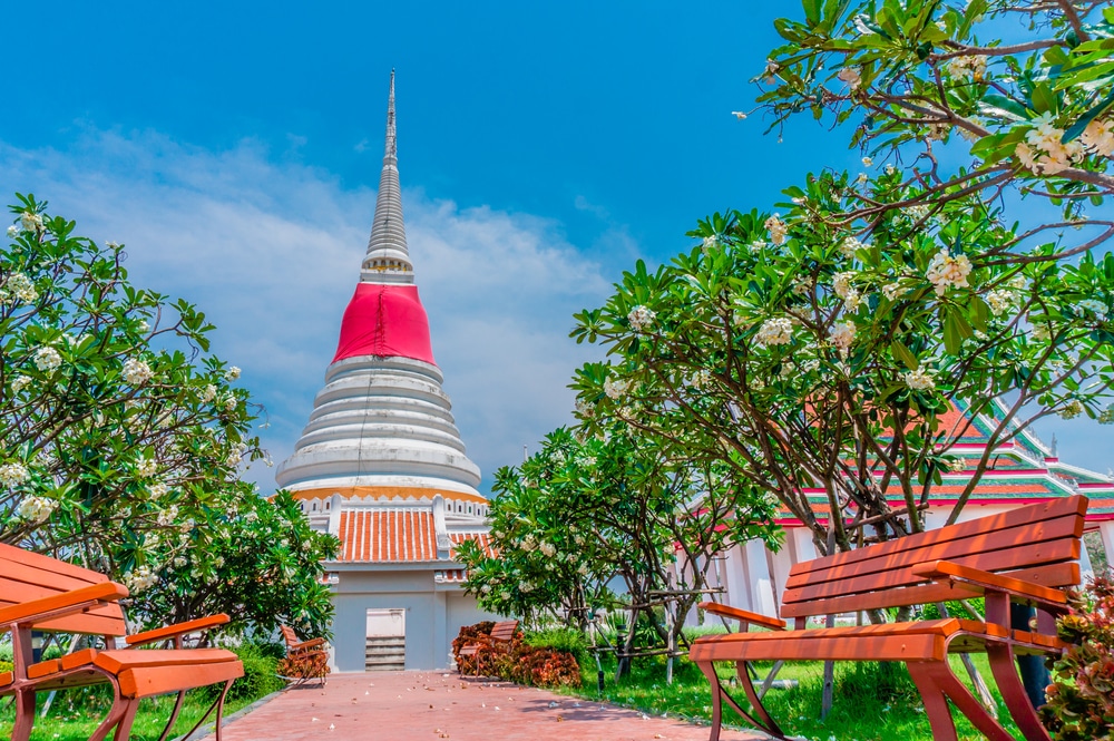 เจดีย์สีขาวและแดงล้อมรอบด้วยต้นไม้ที่บานสะพรั่งและม้านั่งไม้สองตัวใต้ท้องฟ้าสีฟ้าสดใส ถ่ายทอดเสน่ห์อันสงบของ เที่ยวบางปู.