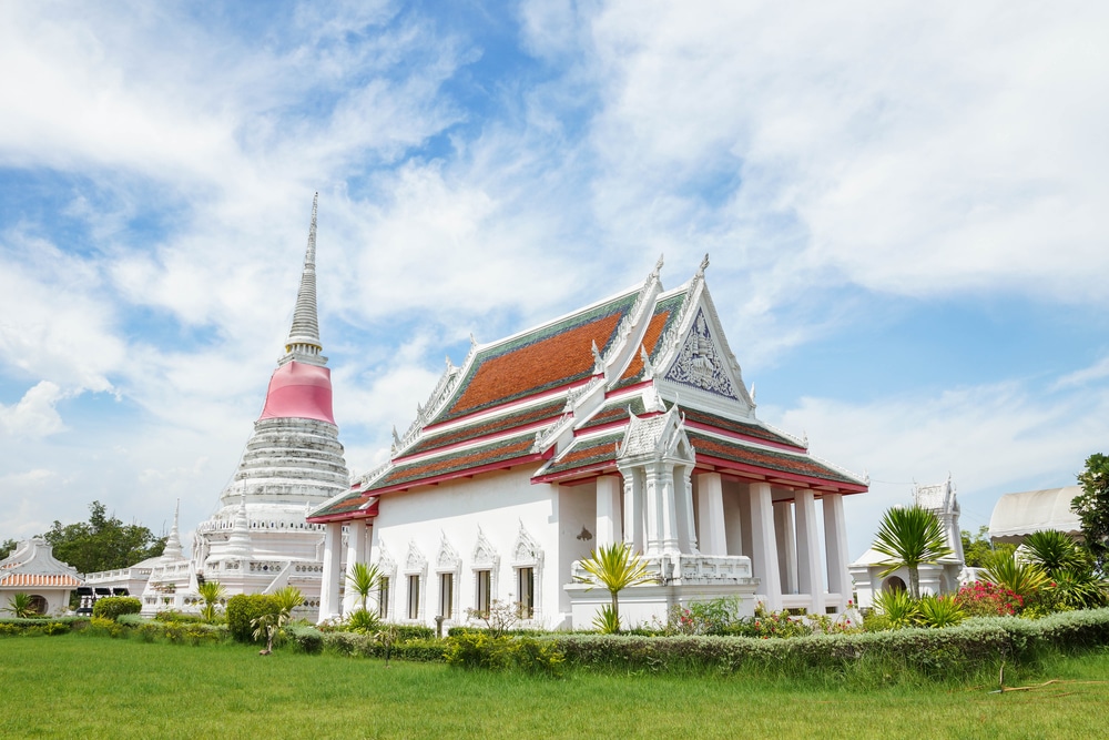 วัดไทยแบบดั้งเดิมที่มีหลังคาซ้อนชั้นสีแดงและขาวตั้งอยู่ข้างสถูปสีขาวที่มีฐานสีชมพู ท่ามกลางท้องฟ้าสีฟ้าสดใสและหญ้าสีเขียว—เป็นภาพที่สงบเหมาะสำหรับผู้ที่มองหาสถานที่ท่องเที่ยวบางปู.