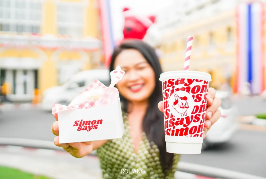 ผู้หญิงคนหนึ่งถือแก้วเครื่องดื่มและกล่องอาหารที่มีตราสินค้า Simon Says Ice Cream อยู่กลางแจ้ง โดยมีอาคารและถนนที่เบลออยู่เบื้องหลัง.