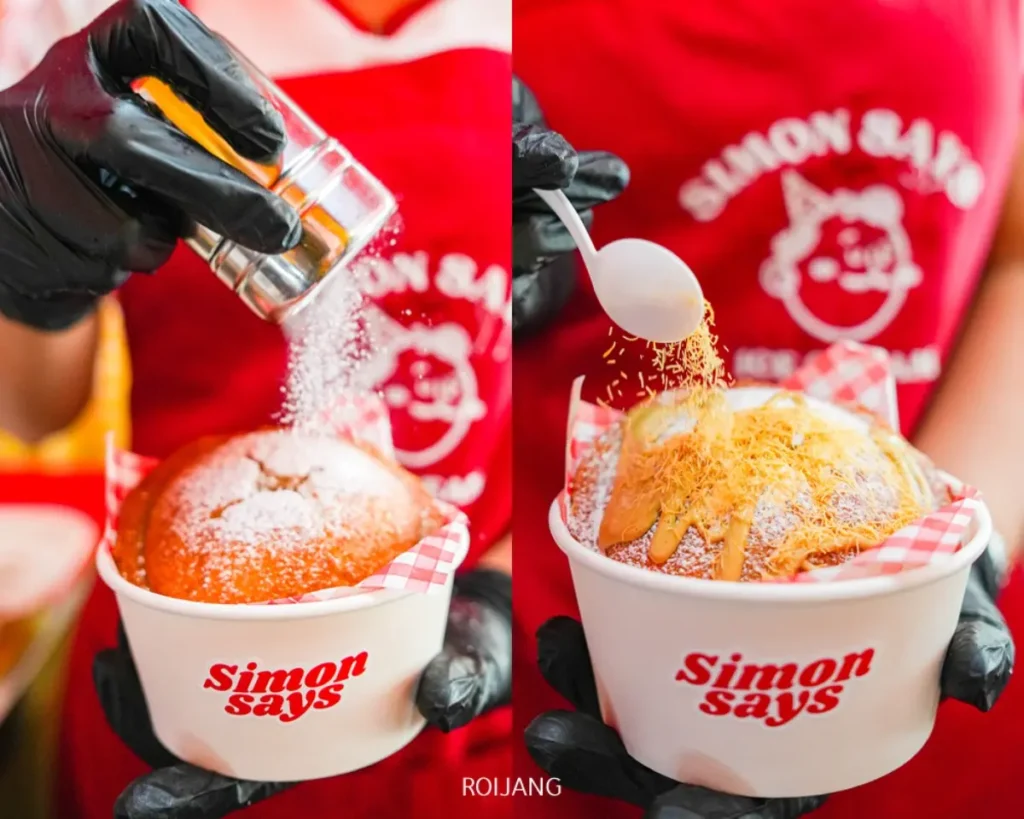 สองมือโปรยน้ำตาลและท็อปปิ้งสีน้ำตาลลงบนขนมอบฟูในชามที่มีแบรนด์ "Simon Says Ice Cream" โดยมีคนในผ้ากันเปื้อนสีแดงอยู่เบื้องหลัง.