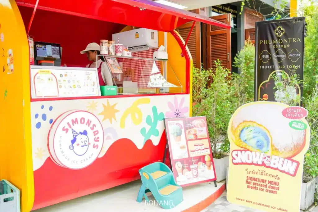บุคคลหนึ่งยืนอยู่ภายในคีออสสีสันสดใสของ Simon Says Ice Cream และของหวาน โดยมีป้ายโฆษณาขนาดใหญ่ของ Snow Bun แสดงอยู่ด้านนอก.