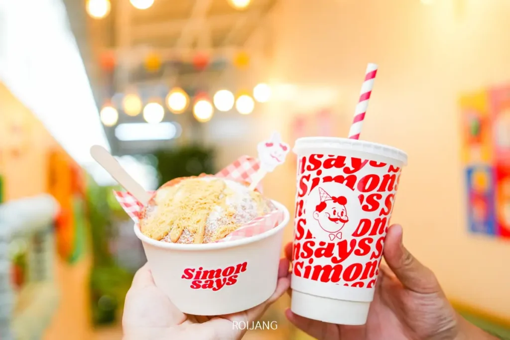 สองมือยกถ้วยของหวานและแก้วกระดาษที่มีหลอดดูดขึ้นมา ทั้งสองมีตรา "Simon Says Ice Cream" ตัวอักษรสีแดง วางอยู่บนพื้นหลังในร่มที่สว่างไสว.