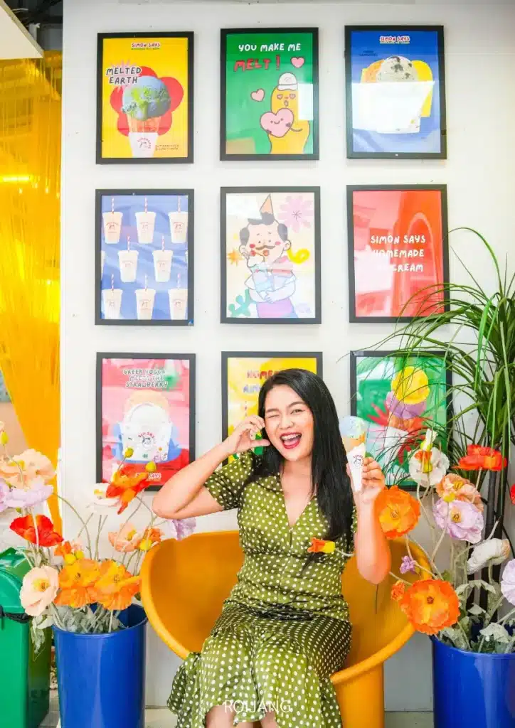 ผู้หญิงคนหนึ่งสวมชุดกระโปรงลายจุดสีเขียว นั่งอยู่บนเก้าอี้สีเหลือง กำลังเพลิดเพลินกับไอศกรีม Simon Says โดยมีดอกไม้หลากสีล้อมรอบ และโปสเตอร์ติดกรอบประดับอยู่บนผนังด้านหลัง.