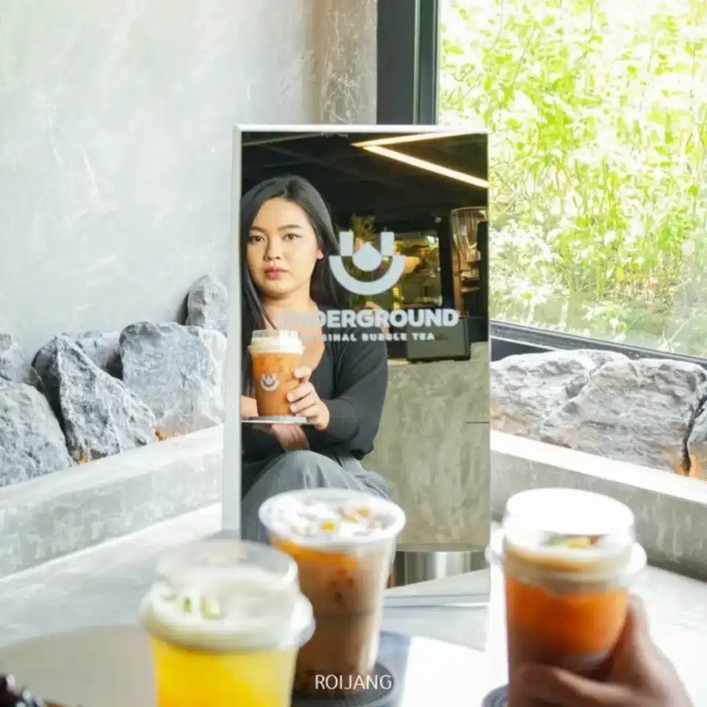 ผู้หญิงถือแก้วชานมไข่มุกสะท้อนในกระจกที่มีข้อความ "UNDERGROUND Official Bubble Tea" ขณะที่เครื่องดื่มที่ได้แรงบันดาลใจจากใต้ดินสามแก้ววางอยู่บนโต๊ะในเบื้องหน้า.