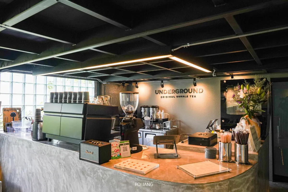 เคาน์เตอร์ร้านชานมไข่มุกใต้ดินสมัยใหม่ มีเครื่องชงกาแฟ แก้ว เครื่องใช้ และเมนู พร้อมป้าย "ชานมไข่มุกต้นตำรับใต้ดิน" บนผนังด้านหลัง.