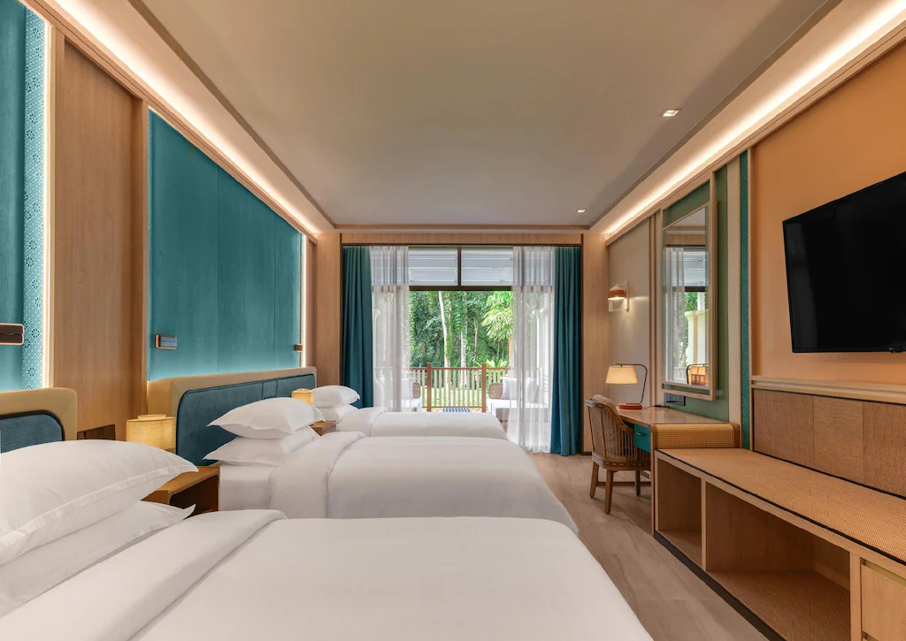 ห้องพักโรงแรมสมัยใหม่พร้อมเตียงสามเตียง โทรทัศน์ติดผนัง โต๊ะและเก้าอี้ หน้าต่างบานใหญ่ และระเบียงที่มองเห็นวิวสีเขียวชอุ่ม—เหมาะสำหรับการพักผ่อนหลังจากวันที่สำรวจสถานที่ท่องเที่ยวในกระบี่.